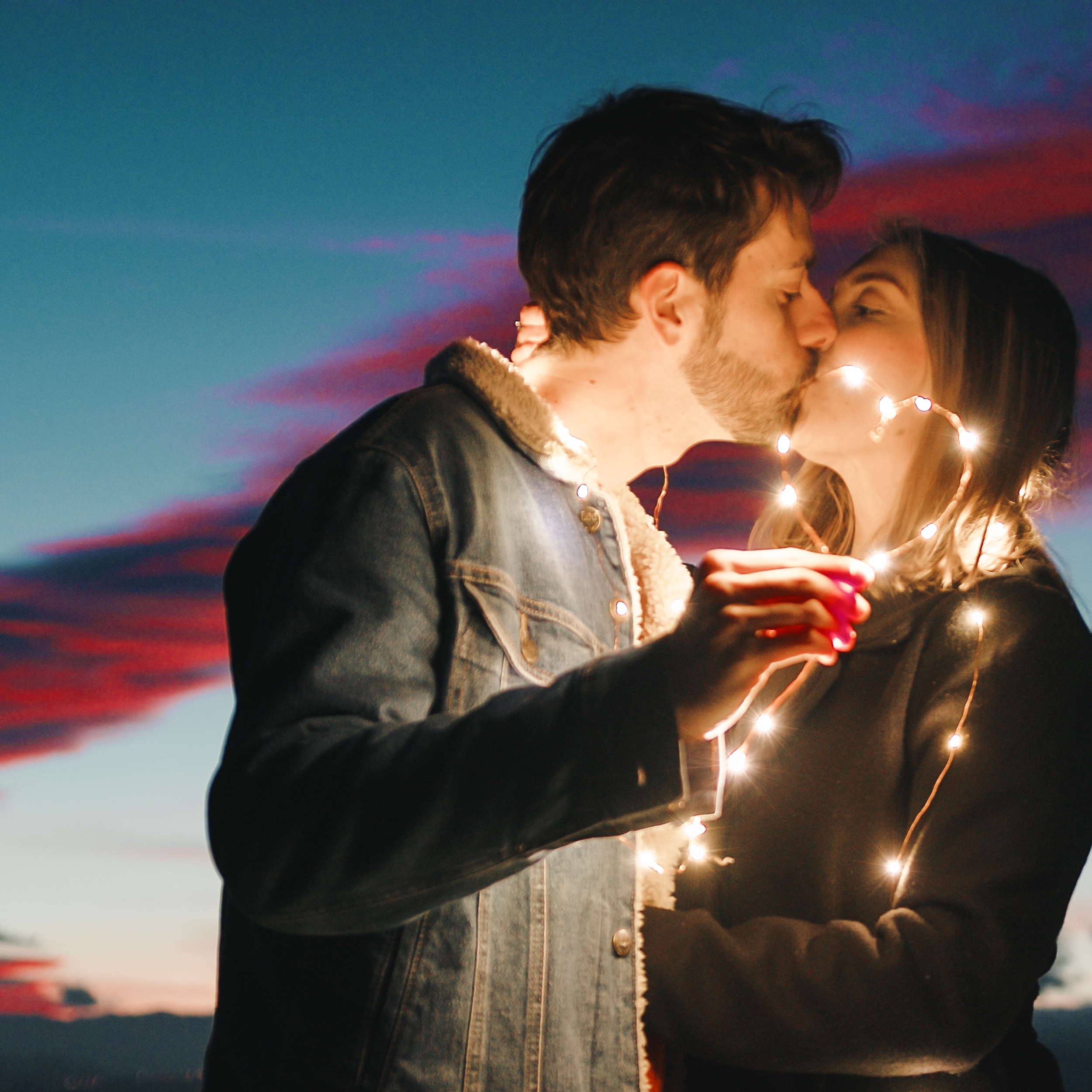 Paar küsst sich mit Lichterkette in den Händen vor dem Abendhimmel